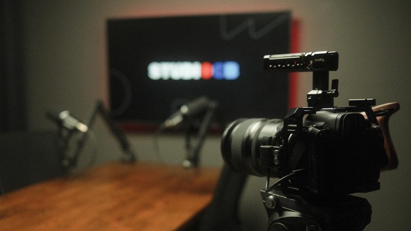 Câmera de vídeo em primeiro plano focada na mesa de podcast configurada para duas pessoas. Equipamento de filmagem profissional.