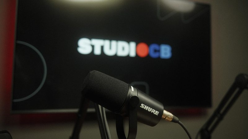 Close em microfone Shure de podcast com fundo desfocado e telão ao fundo. Equipamento de áudio de alta fidelidade.