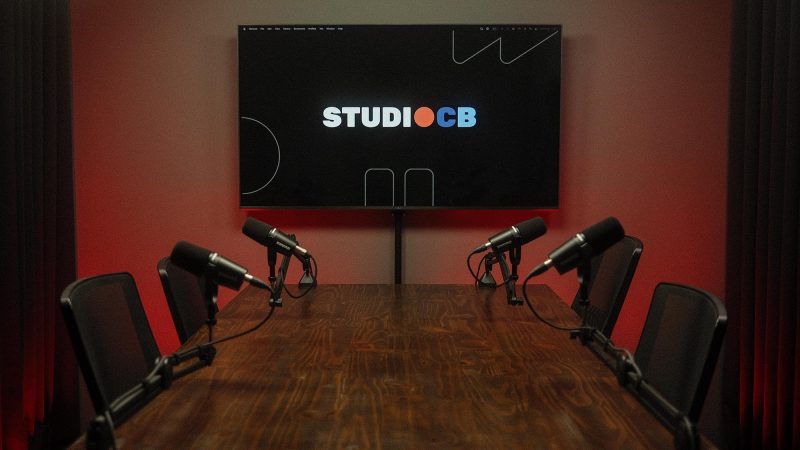 Mesa de podcast vista de frente com 4 cadeiras ergonômicas idênticas e microfones alinhados. Estrutura profissional e simétrica.