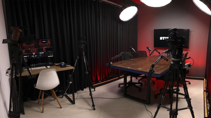 Foto panorâmica do estúdio mostrando a mesa de podcast e a ilha de edição/monitoramento do técnico lado a lado.